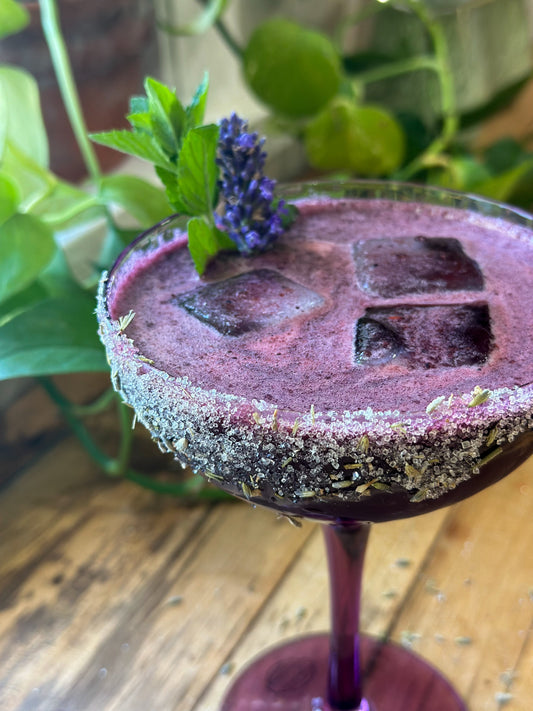 Purple Third Eye Mocktail Elixir in a sugared-rim glass with lavender and mint garnish, served over ice on a wooden table.